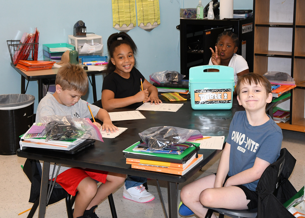 Four children seated at a table in a classroom, with various school supplies and a trash can nearby.