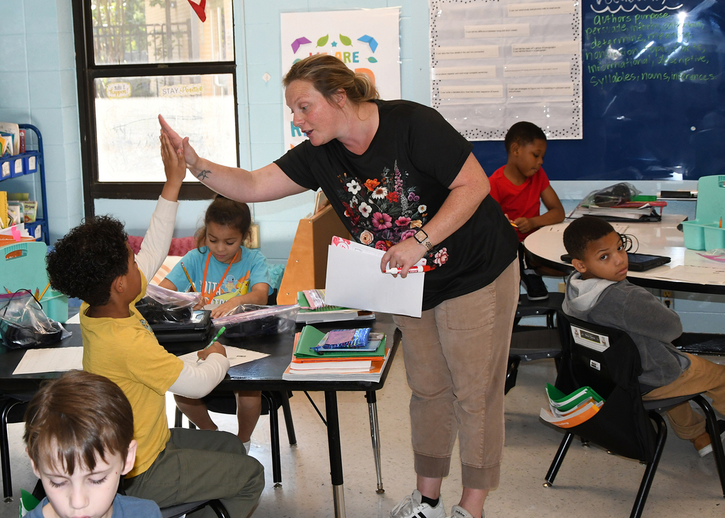 A woman stands in front of students at a table, holding paper, and giving high fives.