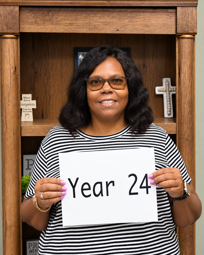 Woman in a striped shirt holding a sign that says "Year 24" in front of a wooden bookshelf.