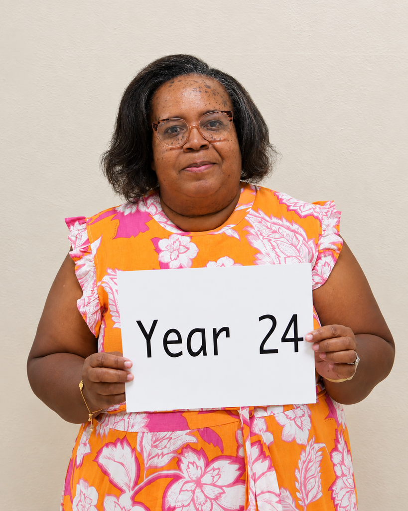 A woman in an orange floral dress holds a white sign with black letters, "Year 24". She is in a room with a light-colored wall.