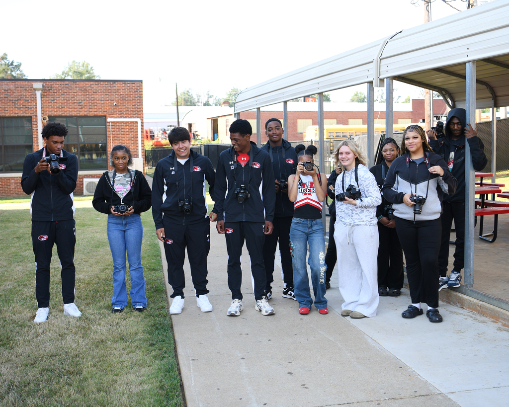 A group of individuals stands on a sidewalk, wearing sports jackets and holding cameras. Behind them, a brick building and a metal canopy are visible.