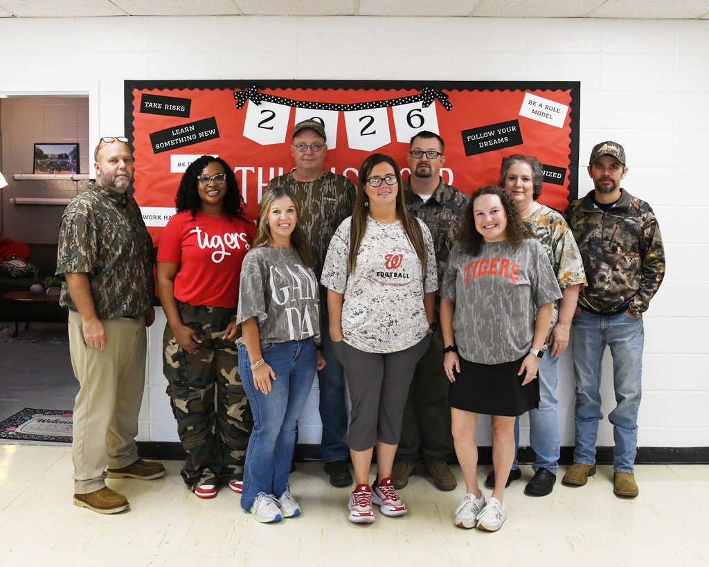 A group of adults wearing sports jerseys and camouflage stand before a bulletin board.