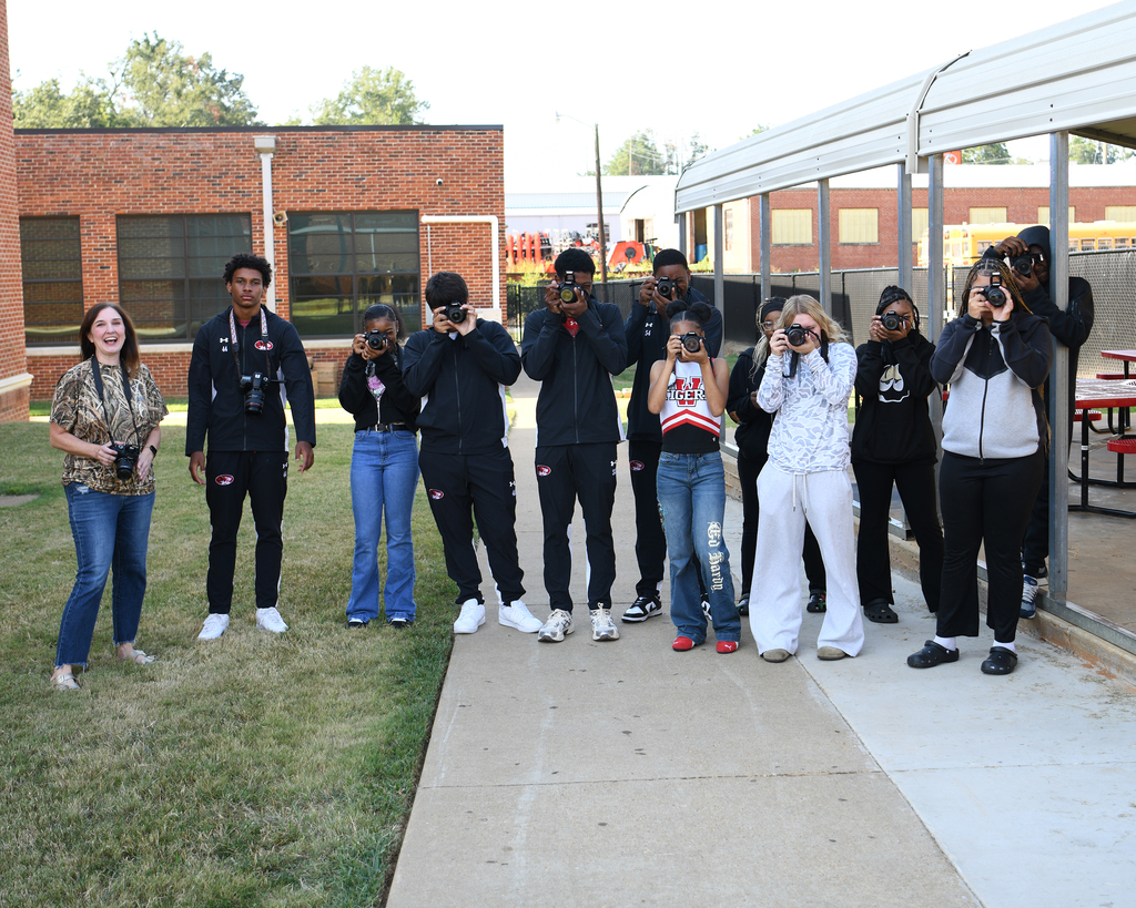 A group of students wearing jackets and holding cameras, posing on a concrete walkway in front of a brick building.