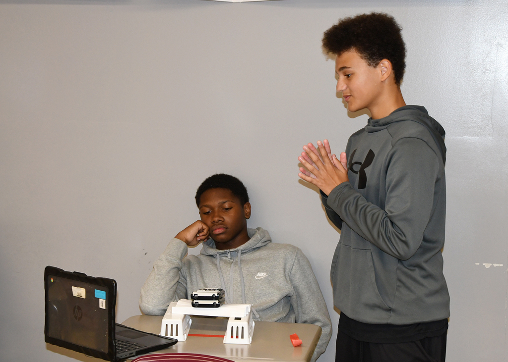 Two people in hoodies, one seated at a table with a laptop, the other standing behind him clapping.