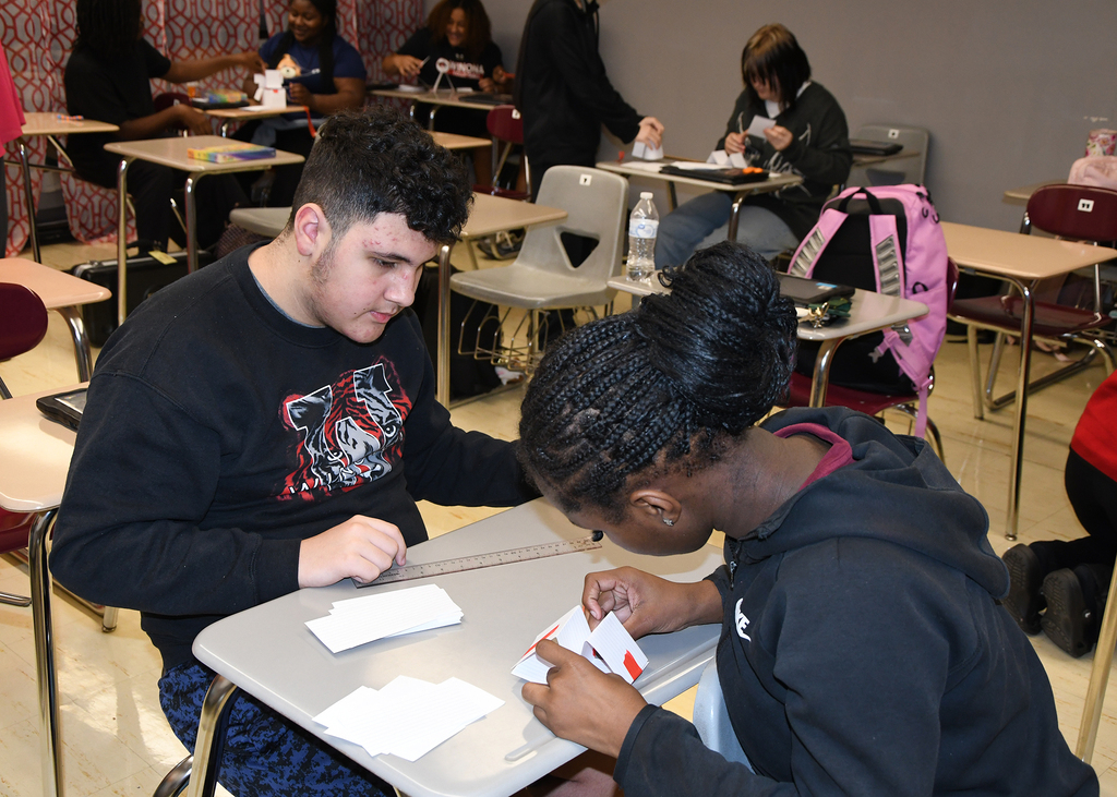 Students sit at desks, with two individuals closely working on papers, surrounded by other students.