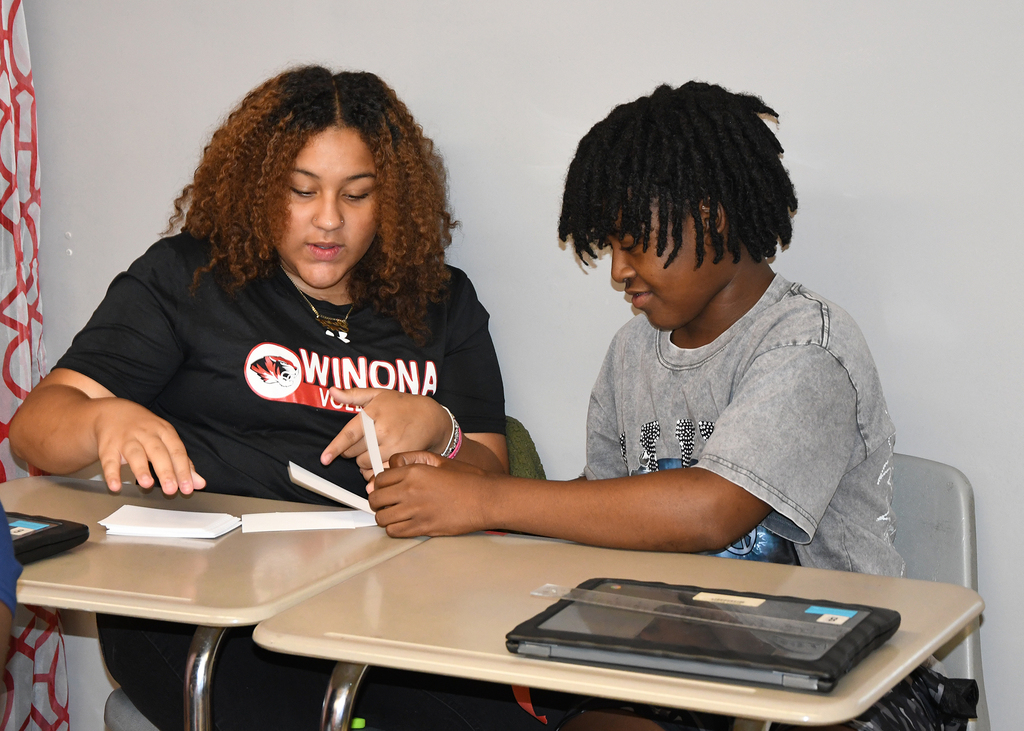 A woman and a boy sit at a desk, engaged in reading. The woman has curly hair and the boy has braided hair.