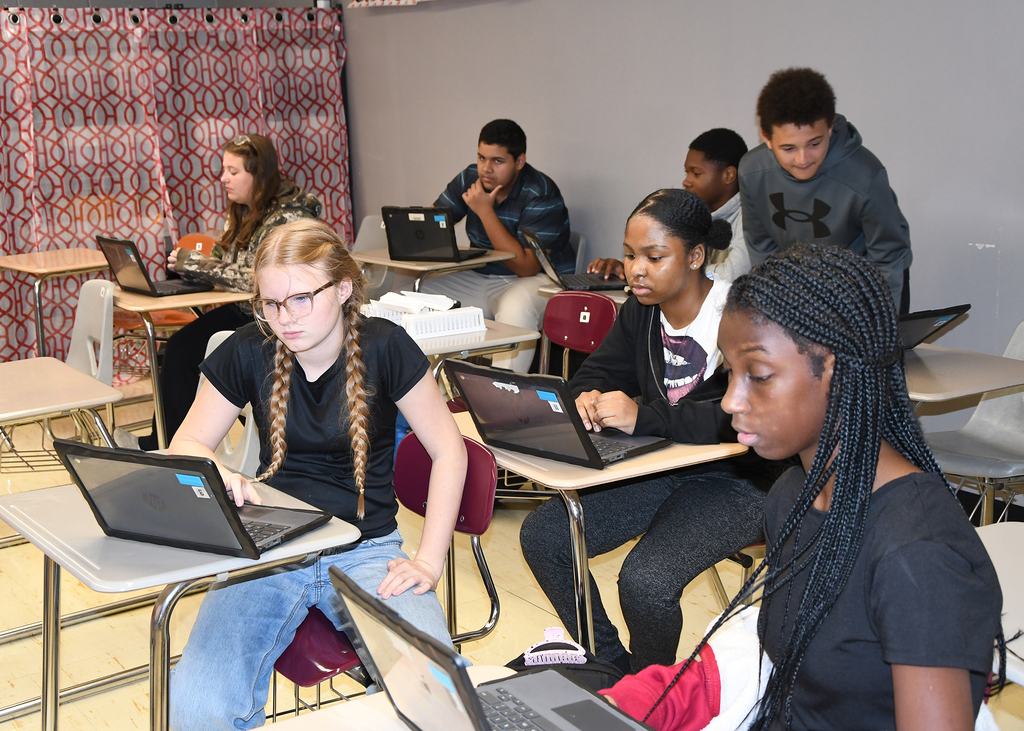 A classroom with several students using laptops on desks. One student has braided hair and sits at the front.