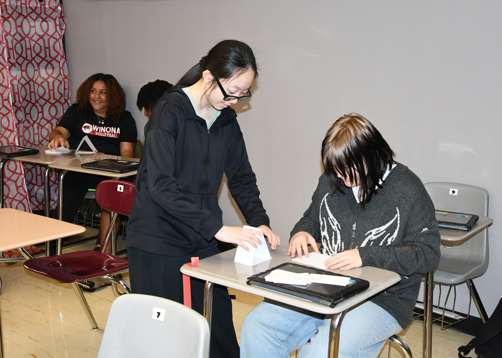 A person standing over a seated person at a desk, both holding paper and a tablet.