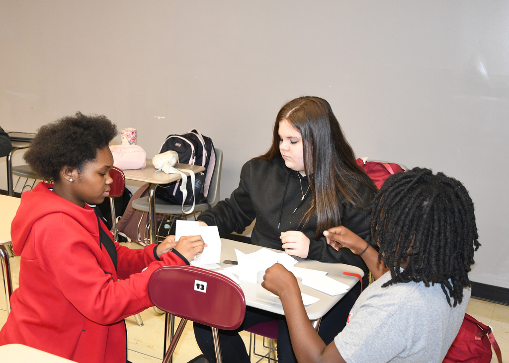 Three students seated at a table, engaged in discussion and holding paper. One student wears a red jacket, another a gray shirt.
