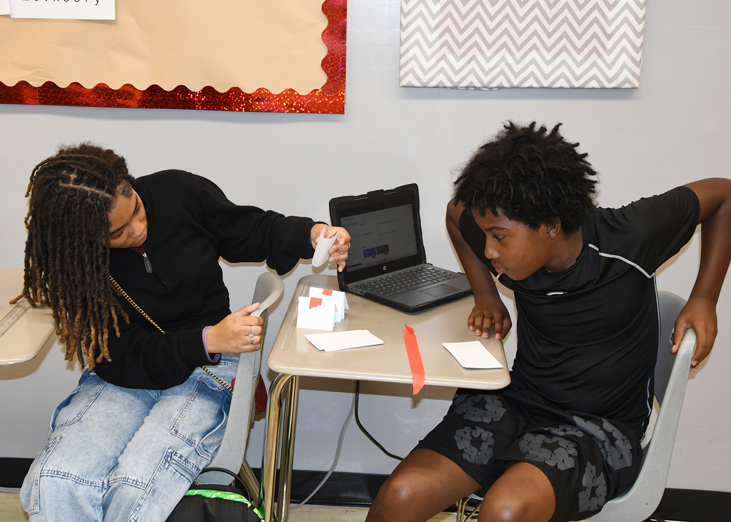 Two students seated, one uses a computer, the other looks at cards. Behind them is a wall with colorful decorations.