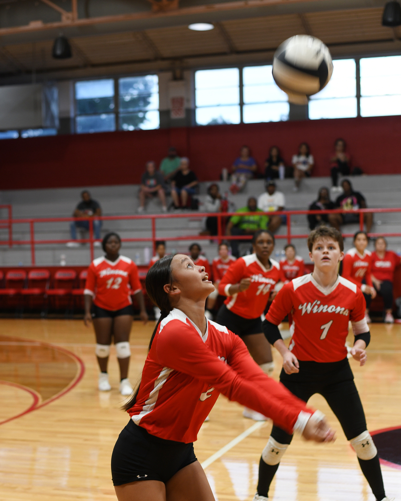 A volleyball game in progress with a player in red and white jersey jumping to hit the ball.