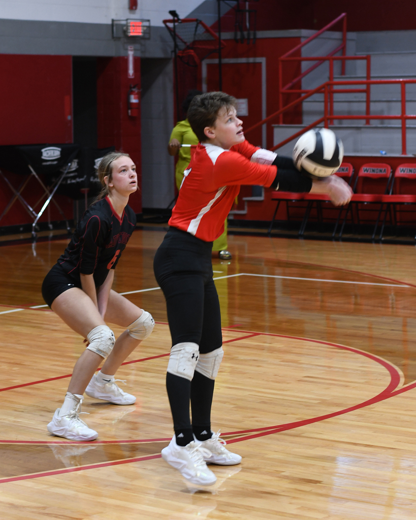 Two girls play volleyball on a court. One girl is in a red and white uniform, the other in black.