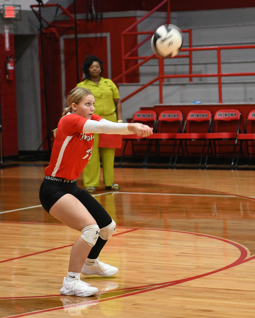 A young woman in red plays volleyball on an indoor court. A woman in green stands behind her near red chairs.