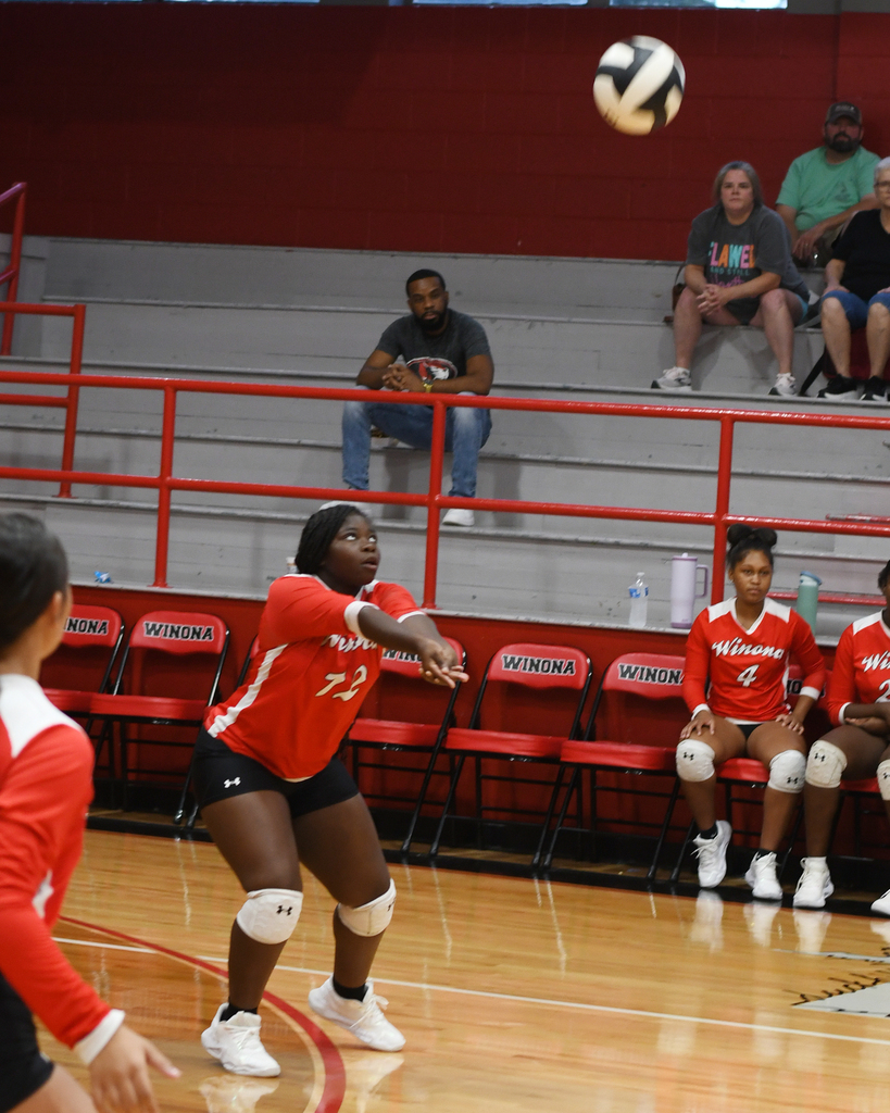 Volleyball in air as player in red jersey prepares to hit it. Spectators sit on red bleachers.