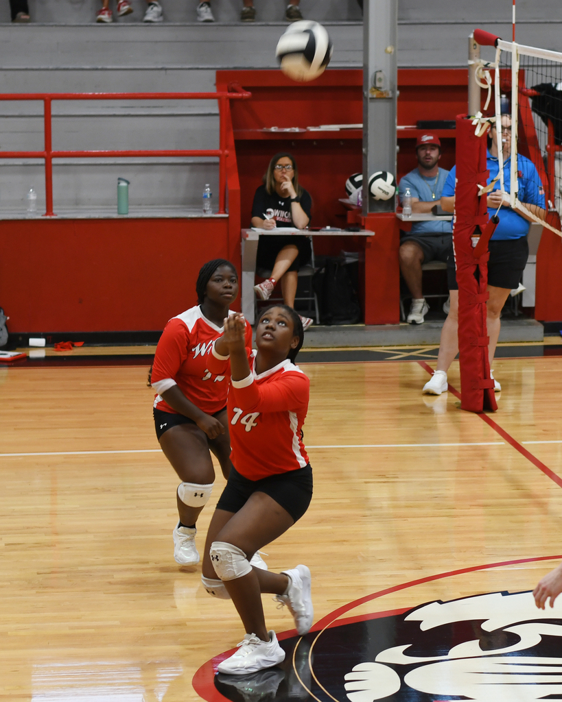 Two players in red jerseys on a court, one jumping to hit a volleyball. Spectators watch from bleachers.