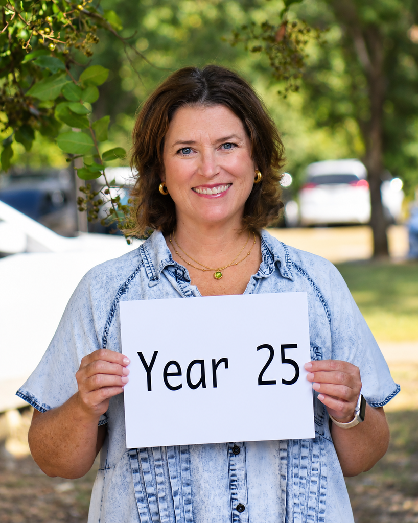 A woman smiling and holding a white sign that reads "Year 25." She is in a park with parked cars and trees in the background.