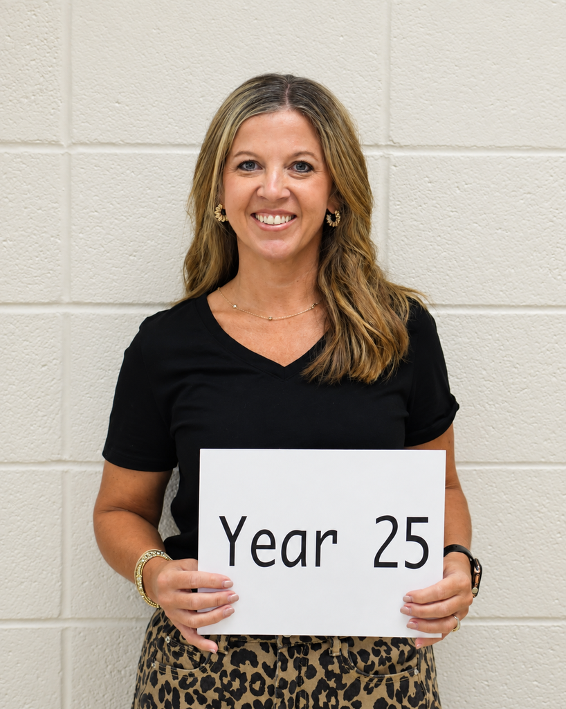 A woman holds a sign that reads "Year 25." She is wearing a black V-neck shirt, leopard-print skirt, and gold jewelry.