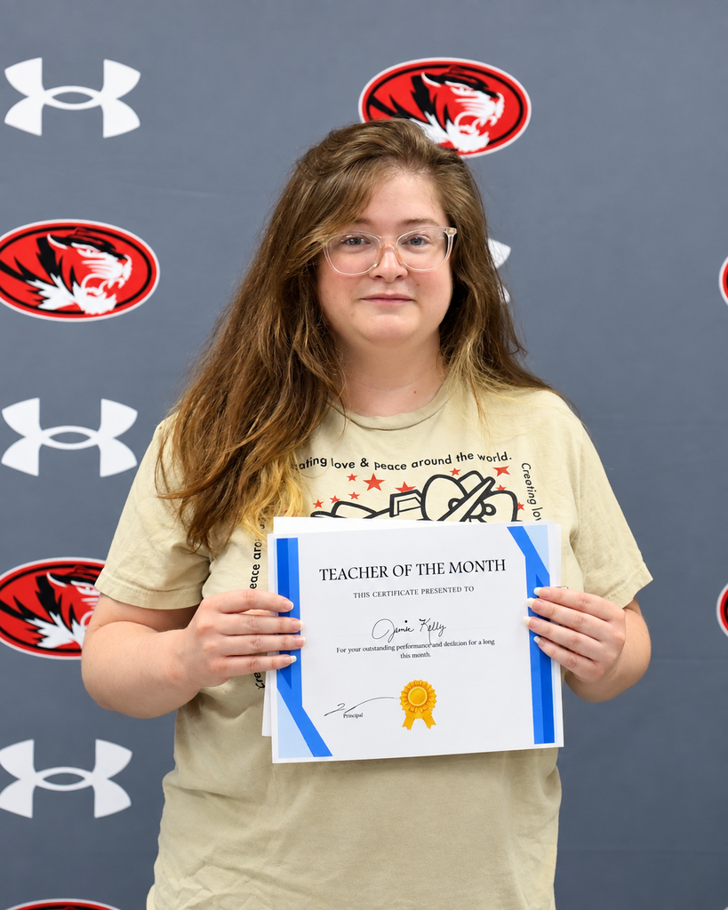 A woman wearing glasses holds a certificate for Teacher of the Month, standing in front of a wall with red tigers.