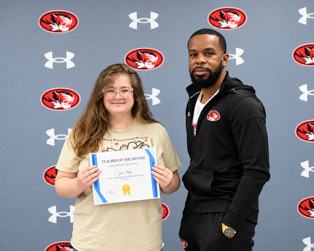 A young woman holds a certificate next to a man in front of a wall with Under Armour logos.