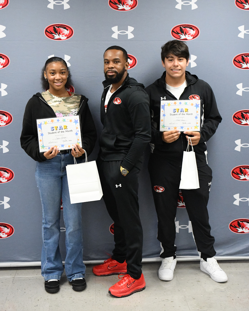 Three individuals stand in front of a branded backdrop. Each holds a certificate. Two men wear black jackets and pants. A woman wears a jacket, jeans, and holds a bag.