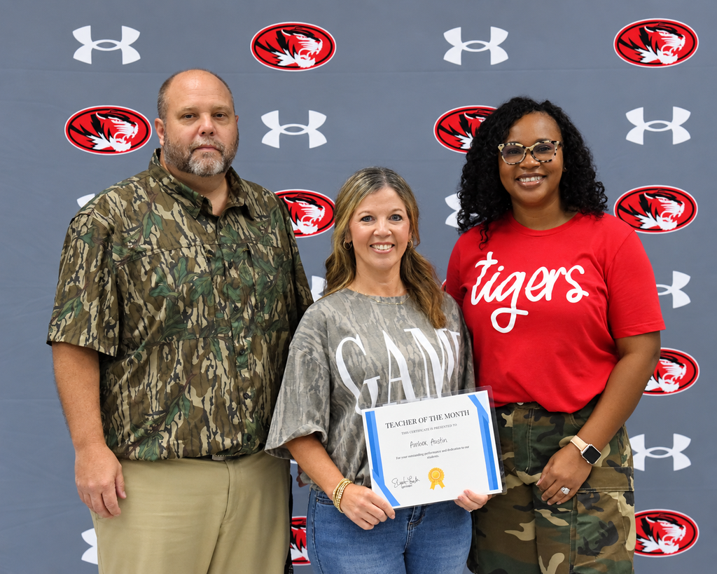 Three adults stand in front of a wall with red logos. A woman holds a certificate, while two others look on.