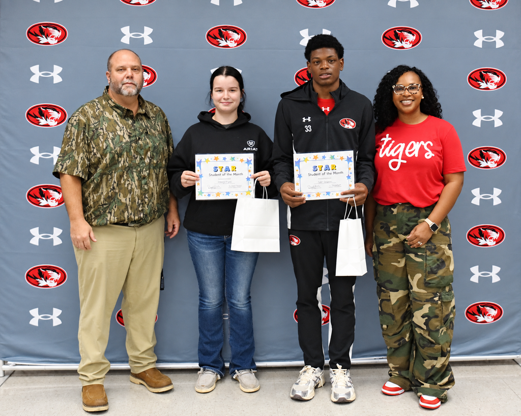 Five individuals stand together, posing for a photo with certificates. Two hold white bags. A blue wall with Under Armour logos and a red tiger logo is in the background.