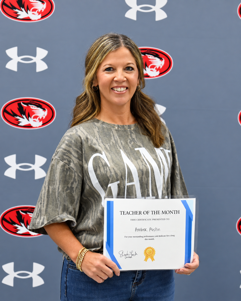 A woman holds a certificate reading "Teacher of the Month" in front of a gray wall with red logos.