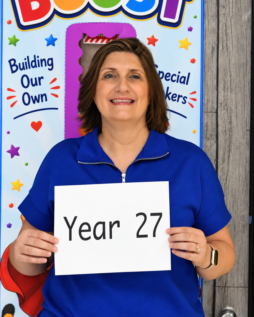 A smiling woman in a blue shirt holds a sign that reads "Year 27" in front of a colorful backdrop with text.