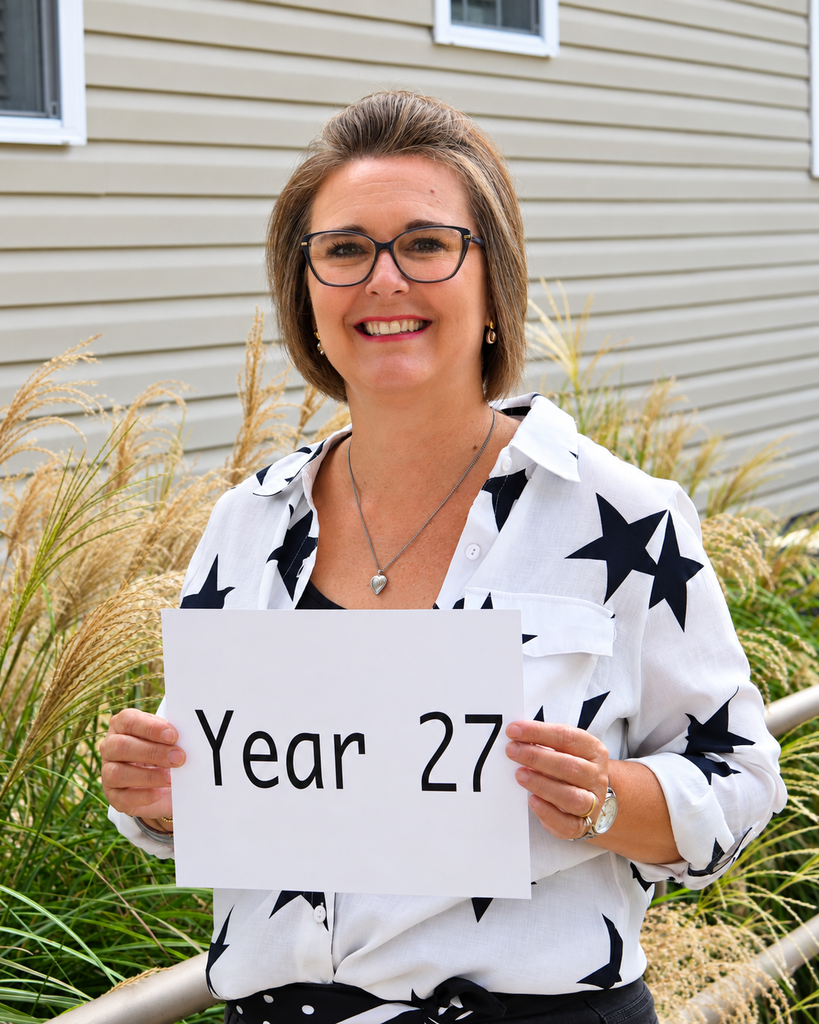 Woman with glasses stands outside a house, holding a sign that says "Year 27." Behind her, there is a grassy area.