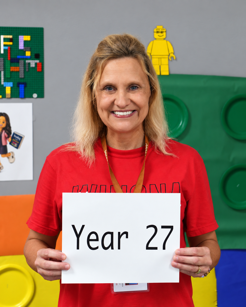 A woman holding a sign that says "Year 27" stands in a room with colorful building blocks.