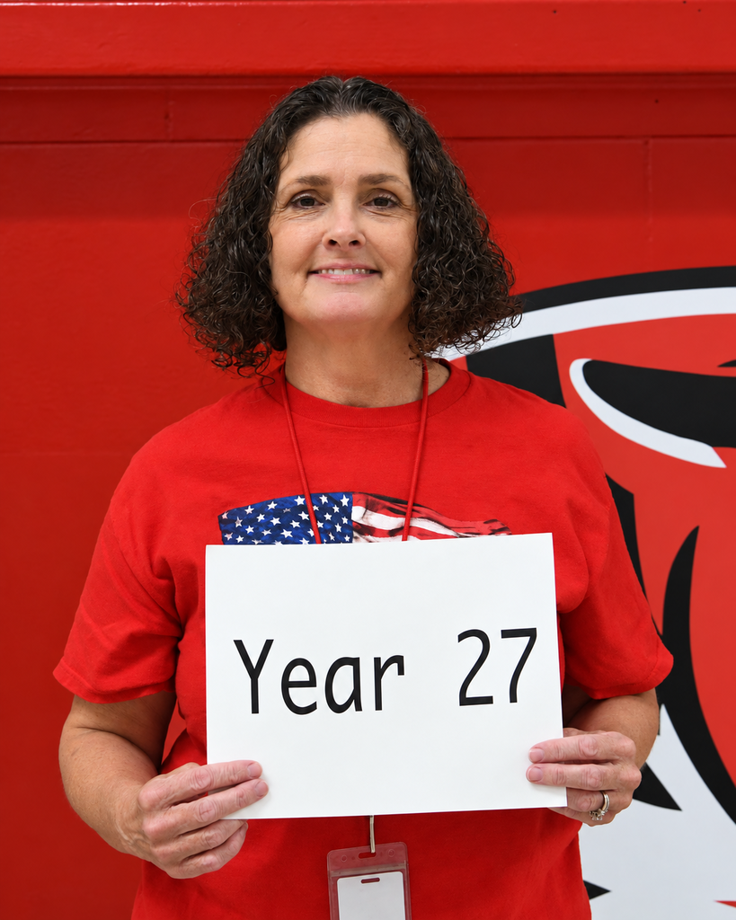 Woman with curly hair smiles in a gym, holding a white sign that says "Year 27." Red background with a large black and white logo.