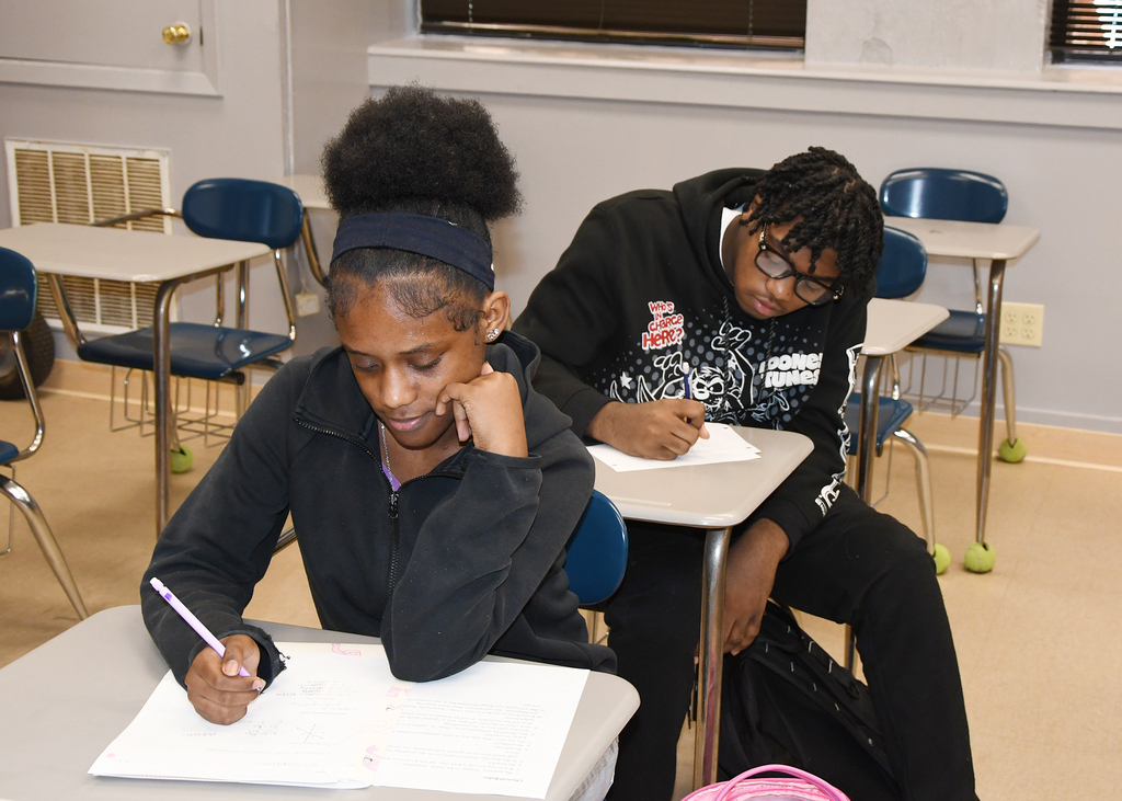 Two students seated at desks in a classroom. One looks at a paper while writing, the other rests head on a desk.