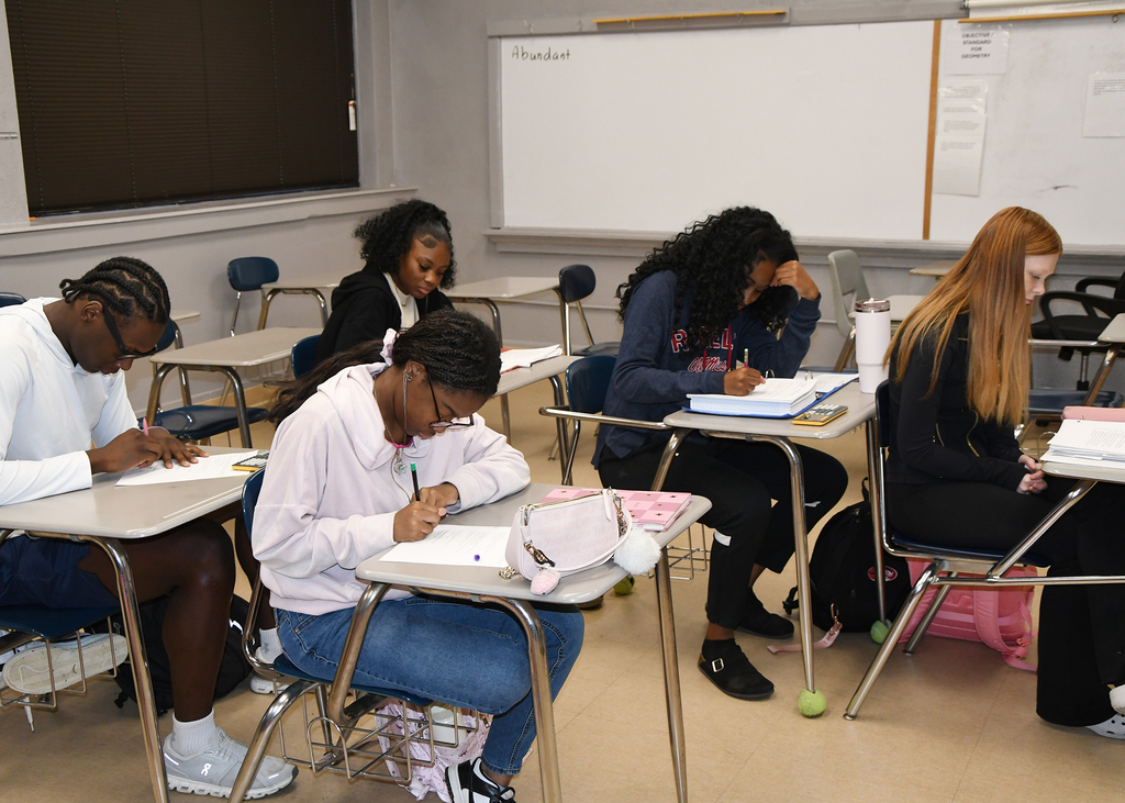 Students seated at desks, writing on paper, one woman with glasses, another with red hair, two other women, and a whiteboard.