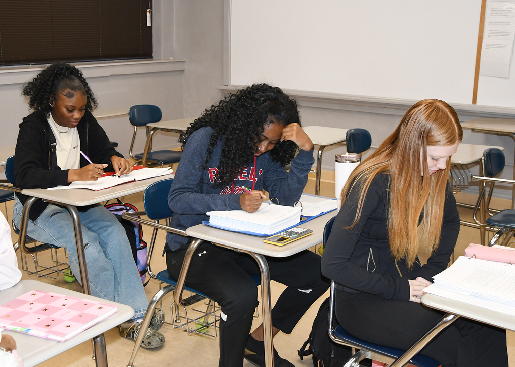 Four students in a classroom. Three sit at desks with books, and one leans on a desk.