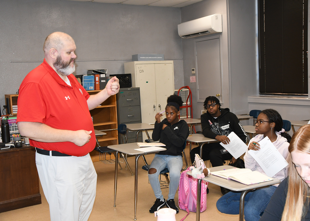 A man stands before a classroom, gesturing as students sit at desks, some with books and papers.