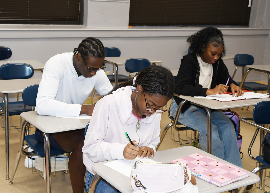 Three students in a classroom, each seated at a desk, writing in notebooks. Desks have papers and bags.