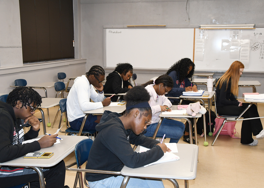 Several students in a classroom, seated at desks, engaged in writing. A whiteboard is mounted on the wall behind them.