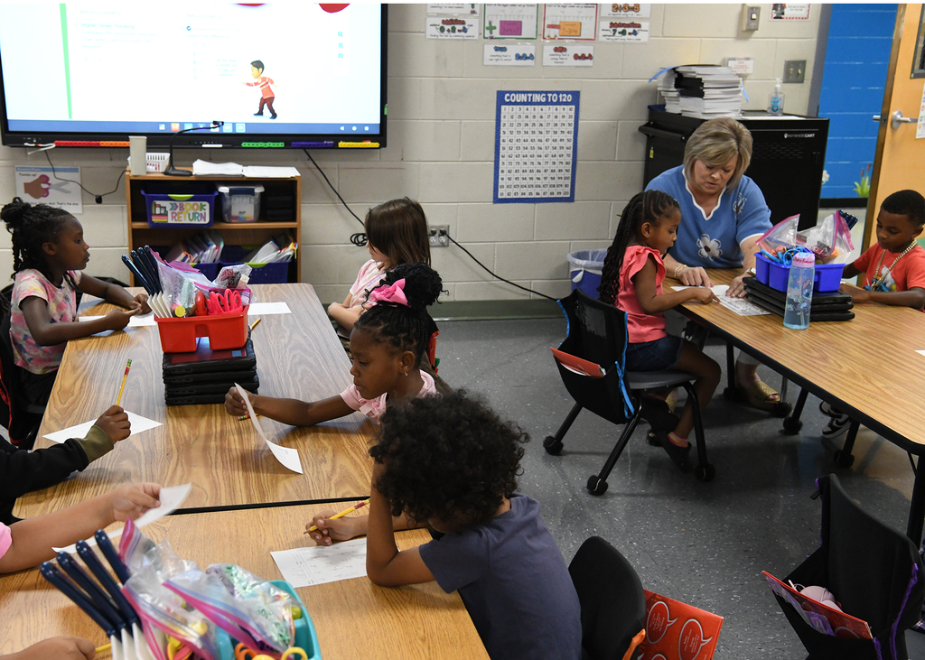 Classroom with children seated at tables, a teacher, and a monitor on a wall.
