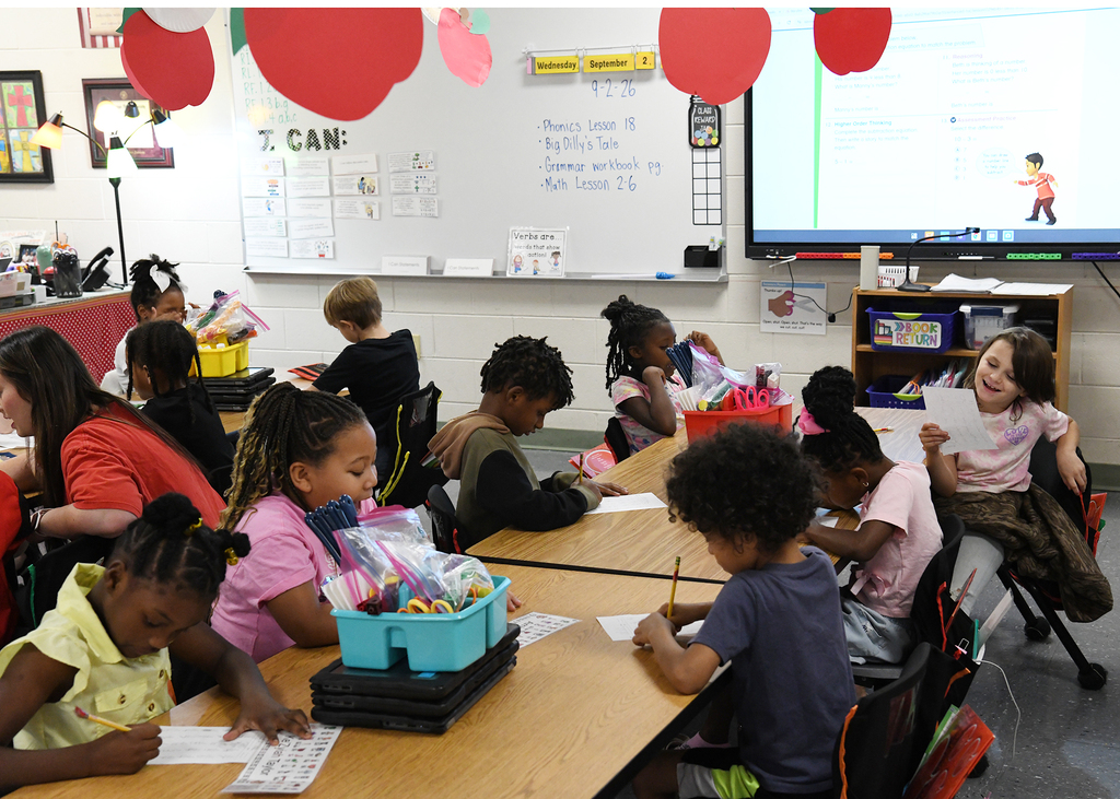 Kids seated at tables in a classroom with a teacher. A monitor and whiteboard behind them.