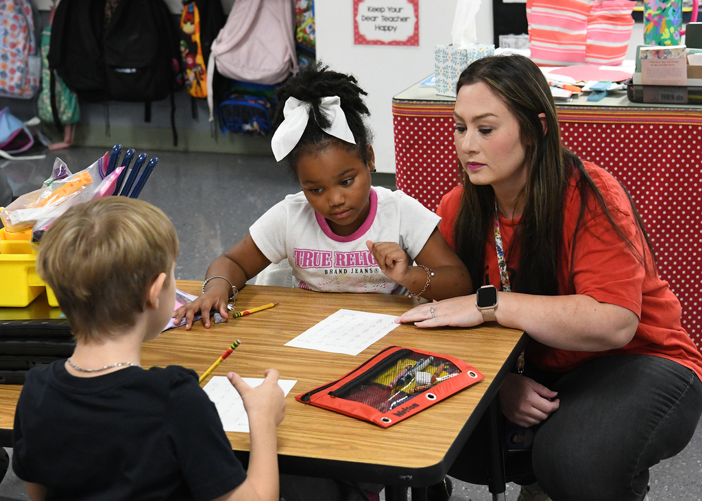 Teacher in red top helping young girl and boy seated at desk. Girl has white bow in hair.