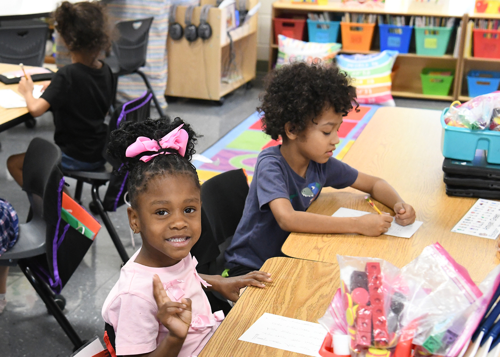 Children seated at desks, one smiling and waving, the other focused on writing, with shelves and colorful bins in background.