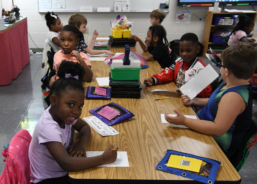 Kids sit around a desk in a classroom, writing and reading. A teacher is present, and various school supplies are on the table.