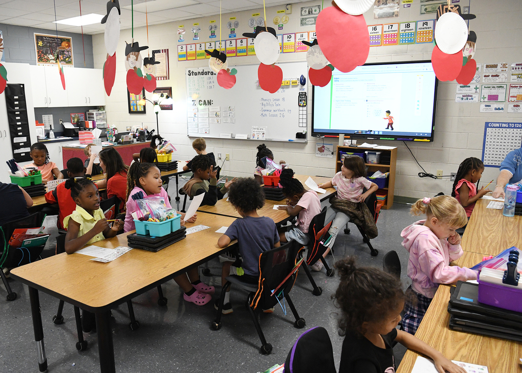 Classroom with children seated at tables; decorations like balloons and apples hang from ceiling; projector screen mounted on wall.