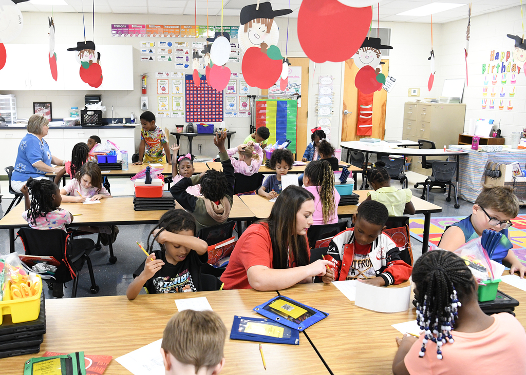A classroom with children seated at desks. A woman helps a child. Various educational materials are on the desks.