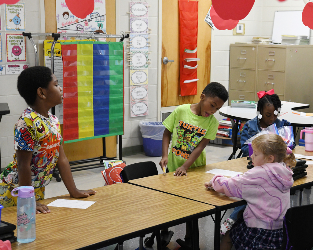 Four kids in a classroom. One stands, another sits, and two write on papers at tables. Rainbow banner, red balloons.