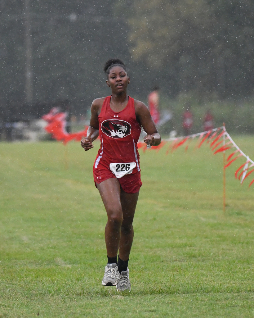 An athlete runs on a grass field during a race in the rain. She wears a red uniform with "226" on it.