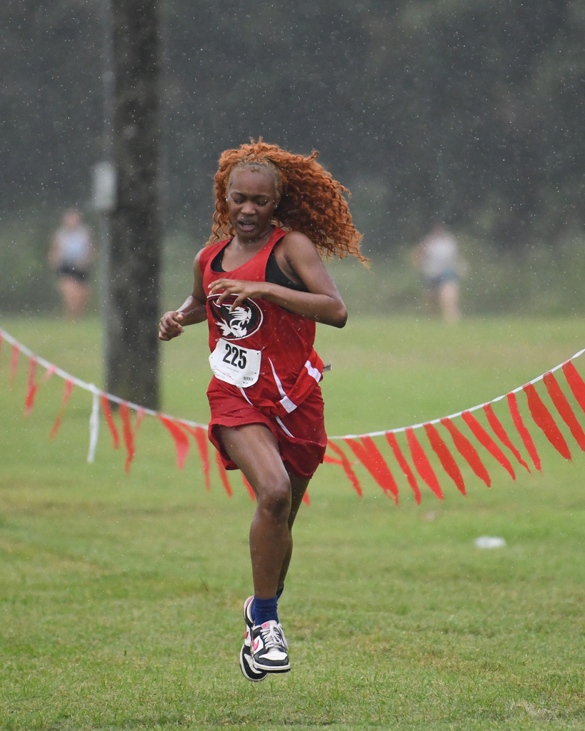 A runner with red hair sprints forward in a red uniform, number 215, on a grassy field with rain.