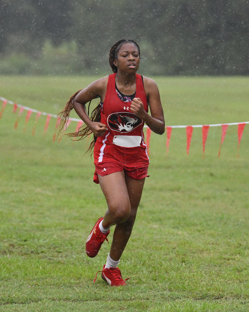 An athlete runs in a cross-country race on a grassy field. She wears a red uniform with white accents and red shoes.