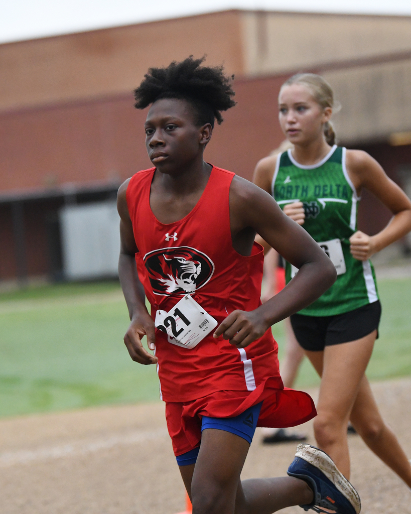 A runner in red and blue shorts with a number 21 bib sprints on a track. A runner in green and black follows behind.