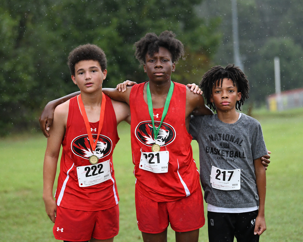 Three young boys with medals stand on a grassy field. Two wear red uniforms, one with the number 21. The third wears a gray shirt.